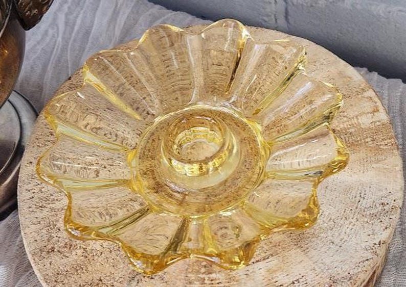 May include: A translucent yellow glass candle holder featuring a scalloped, flower-like edge and a central circular opening designed to hold a taper candle. The glass has a smooth, reflective surface with a distinct radial pattern radiating outward.