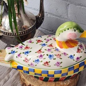 May include: A ceramic duck-shaped container with a lid, featuring a white base adorned with colorful floral patterns. The duck has a green head, pink cheeks, and a yellow beak. The base has a checkered pattern.