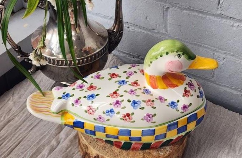May include: A ceramic duck-shaped container with a lid, featuring a white base adorned with colorful floral patterns. The duck has a green head, pink cheeks, and a yellow beak. The base has a checkered pattern.