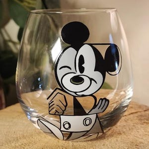Puede incluir: Una copa de vino sin tallo transparente con un dise&ntilde;o de dibujos animados en blanco, negro y gris de Mickey Mouse. La copa est&aacute; sobre una superficie de madera. El personaje de dibujos animados est&aacute; gui&ntilde;ando un ojo.