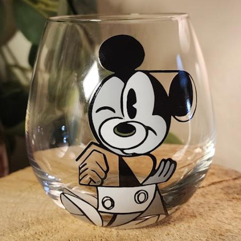 Puede incluir: Una copa de vino sin tallo transparente con un dise&ntilde;o de dibujos animados en blanco, negro y gris de Mickey Mouse. La copa est&aacute; sobre una superficie de madera. El personaje de dibujos animados est&aacute; gui&ntilde;ando un ojo.