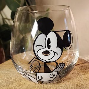 Puede incluir: Una copa de vino sin tallo transparente con un dise&ntilde;o de personaje de dibujos animados. El personaje est&aacute; en negro, blanco y beige, gui&ntilde;ando un ojo y sonriendo. La copa est&aacute; sobre una superficie de madera.