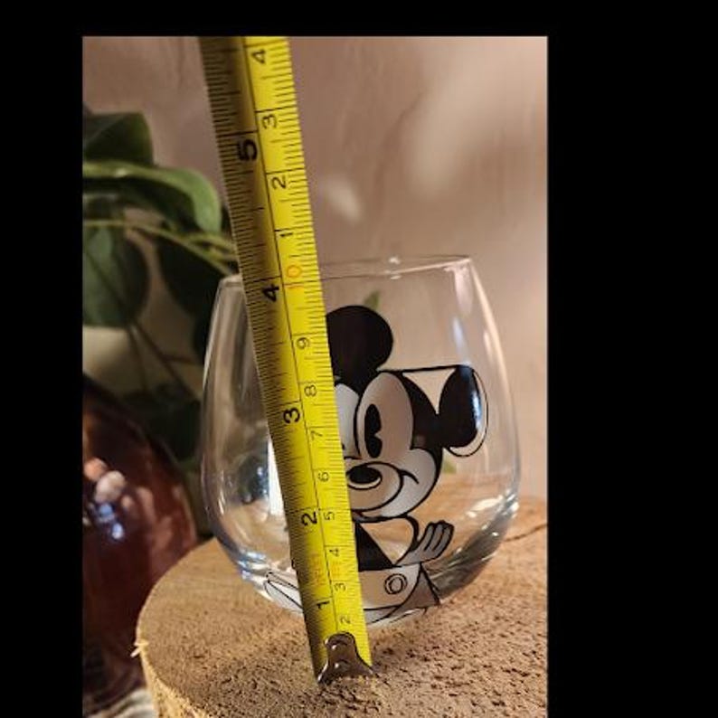 Puede incluir: Una copa de vino sin pie transparente con un dise&ntilde;o de Mickey Mouse en blanco y negro. La copa mide aproximadamente 10 cm de alto, medido con una cinta m&eacute;trica amarilla. La copa est&aacute; sobre una superficie de madera.