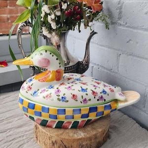May include: A ceramic duck-shaped container with a lid, decorated with colorful floral and checkered patterns. The duck has a green head, pink cheeks, and a yellow beak. It sits on a wooden base, with a floral arrangement in the background.