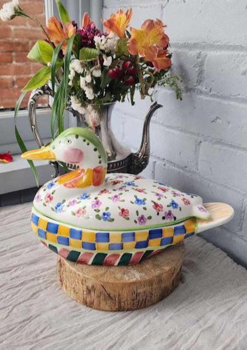 May include: A ceramic duck-shaped container with a lid, decorated with colorful floral and checkered patterns. The duck has a green head, pink cheeks, and a yellow beak. It sits on a wooden base, with a floral arrangement in the background.