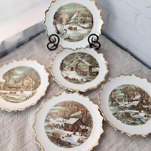 May include: A set of five white ceramic plates with scalloped edges and gold trim. Each plate features a detailed winter landscape illustration with snow-covered cabins, trees, and figures. The text "IN THE WILDERNESS" is printed on the plates.