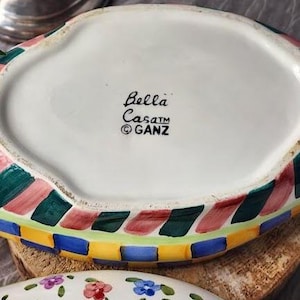 May include: A white ceramic serving dish with a colorful, geometric border in red, green, blue, and yellow. The dish has the text "Bella Casa" and the GANZ logo. The bottom of the dish is visible, along with a floral patterned dish.