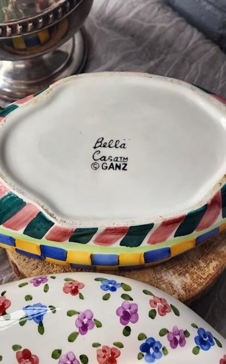 May include: A white ceramic serving dish with a colorful, geometric border in red, green, blue, and yellow. The dish has the text "Bella Casa" and the GANZ logo. The bottom of the dish is visible, along with a floral patterned dish.
