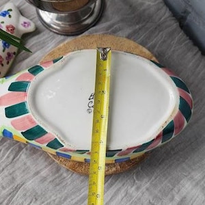 May include: A colorful, oval-shaped ceramic dish with a white interior and a patterned border of green, pink, and blue squares. A yellow measuring tape shows the dish is approximately 6 inches long. The dish sits on a round, cork base.