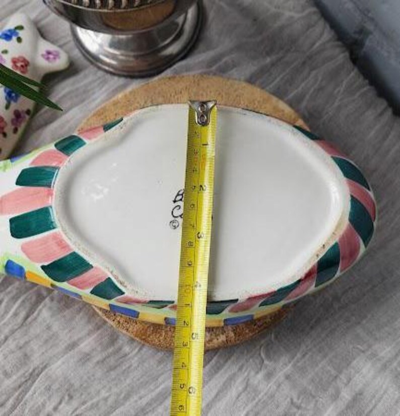 May include: A colorful, oval-shaped ceramic dish with a white interior and a patterned border of green, pink, and blue squares. A yellow measuring tape shows the dish is approximately 6 inches long. The dish sits on a round, cork base.