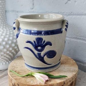 May include: A beige ceramic pot with blue floral designs and two handles. The pot has a blue band around the top and bottom. It sits on a wooden round. A pink and white flower lies on the wood.