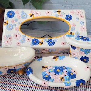 May include: A ceramic bathroom set featuring a soap dish, toothbrush holder, tissue box, and a small bathtub-shaped dish. The set is decorated with blue and pink floral designs on a white background. The bathtub dish is approximately 6 inches long.