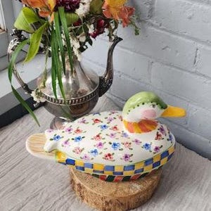 May include: A ceramic duck-shaped trinket box with a floral pattern on the top. The duck has a green head, pink cheeks, and a yellow beak. The base has a checkered pattern in blue, yellow, and red. The box is displayed on a wooden slice.