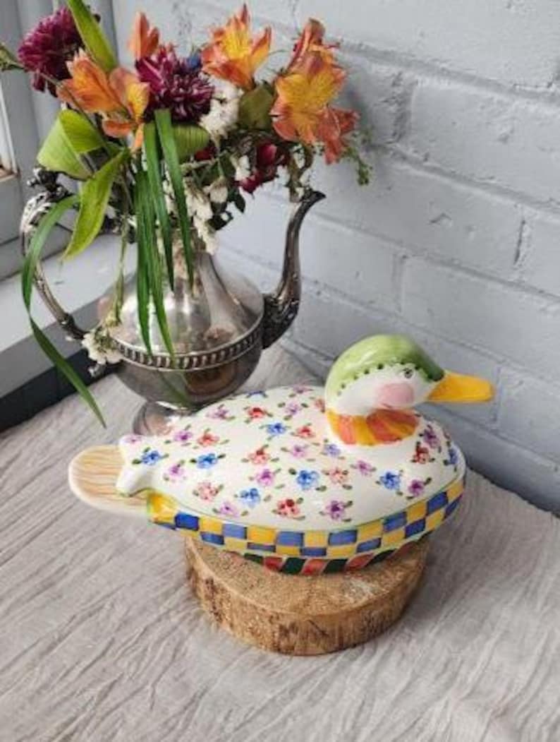 May include: A ceramic duck-shaped trinket box with a floral pattern on the top. The duck has a green head, pink cheeks, and a yellow beak. The base has a checkered pattern in blue, yellow, and red. The box is displayed on a wooden slice.