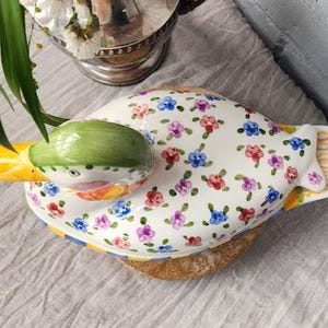 May include: A ceramic duck-shaped dish with a green head and yellow beak. The white body is decorated with small, colorful floral patterns. The dish rests on a wooden base, with a striped tail. A unique decorative piece.