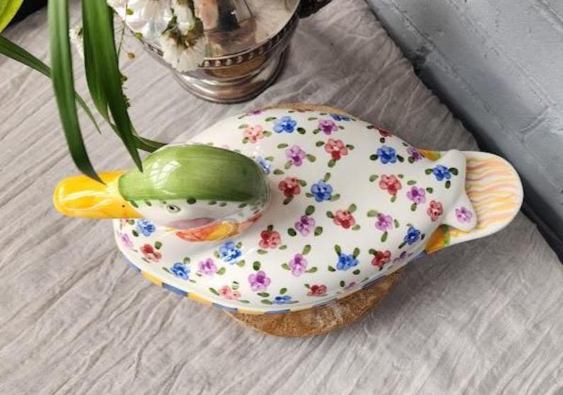 May include: A ceramic duck-shaped dish with a green head and yellow beak. The white body is decorated with small, colorful floral patterns. The dish rests on a wooden base, with a striped tail. A unique decorative piece.