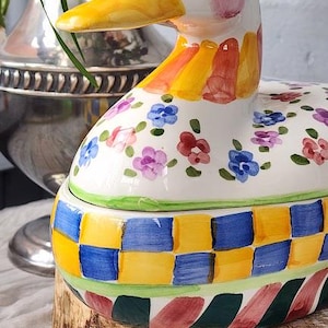 May include: A hand-painted ceramic duck-shaped container. The duck has a yellow beak, green head with black dots, and a white body decorated with colorful flowers. The base features a checkered pattern of blue, yellow, and green.