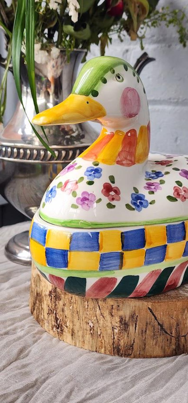 May include: A hand-painted ceramic duck-shaped container. The duck has a yellow beak, green head with black dots, and a white body decorated with colorful flowers. The base features a checkered pattern of blue, yellow, and green.