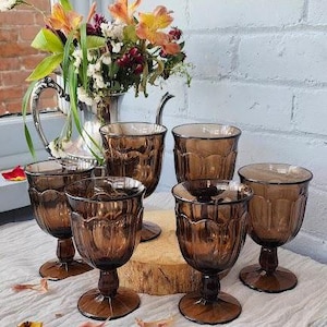May include: Six brown-tinted glass goblets with a tulip-shaped design are arranged on a white surface. A silver pitcher with a bouquet of flowers is in the background. The goblets are on a wooden base.