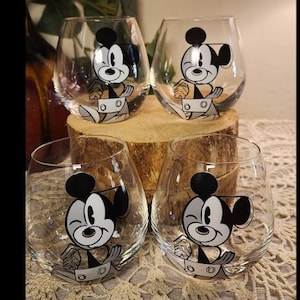 Puede incluir: Cuatro copas de vino sin tallo con un dise&ntilde;o de dibujos animados de Mickey Mouse. Cada vaso transparente presenta una ilustraci&oacute;n en blanco y negro de Mickey Mouse gui&ntilde;ando un ojo. Las copas est&aacute;n dispuestas sobre una base de madera y un mantel de encaje color crema.