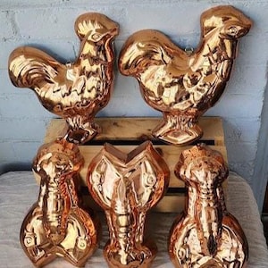 May include: A collection of copper-colored metal molds in the shapes of roosters and abstract figures. The molds are displayed on a wooden crate and a white surface, with a light blue brick wall in the background. The molds are reflective and have detailed features.
