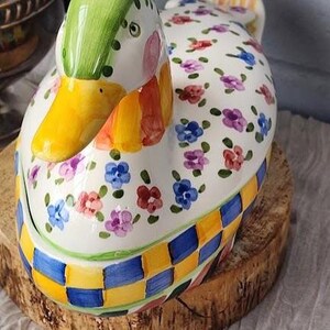May include: A ceramic duck-shaped container with a green head, yellow beak, and floral pattern on a white body. The base features a blue and yellow checkered design. It sits on a wooden slice.