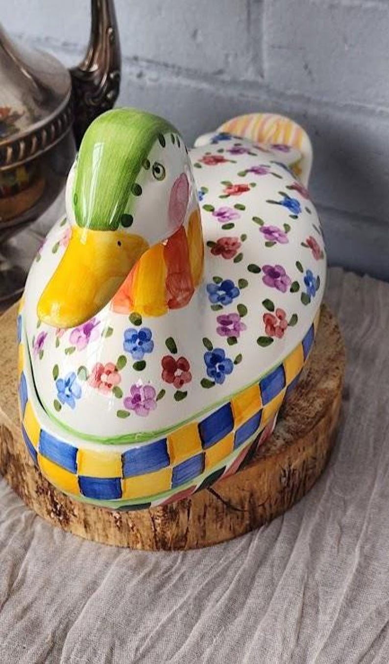 May include: A ceramic duck-shaped container with a green head, yellow beak, and floral pattern on a white body. The base features a blue and yellow checkered design. It sits on a wooden slice.