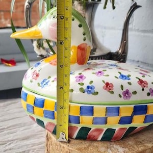 May include: A ceramic duck-shaped container, approximately 5 inches tall, with a lid decorated with floral patterns. The base features a colorful woven design. The duck's head is green, pink, and yellow. A yellow measuring tape is placed next to the container.