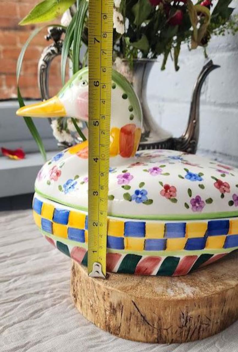 May include: A ceramic duck-shaped container, approximately 5 inches tall, with a lid decorated with floral patterns. The base features a colorful woven design. The duck's head is green, pink, and yellow. A yellow measuring tape is placed next to the container.