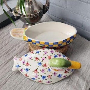 May include: A white ceramic serving dish with a checkered blue and yellow rim and a handle with a painted brush design. Below is a matching floral-patterned dish with a green and yellow handle. The dishes are on a light gray surface.