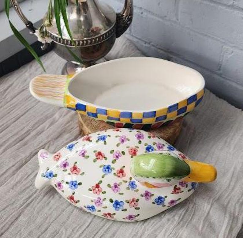 May include: A white ceramic serving dish with a checkered blue and yellow rim and a handle with a painted brush design. Below is a matching floral-patterned dish with a green and yellow handle. The dishes are on a light gray surface.
