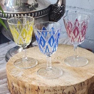 Puede incluir: Tres copas de vino con diseños de diamantes en amarillo, azul y rojo. Se exhiben sobre una superficie de madera, con una jarra plateada al fondo. Diseño de diamantes.