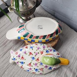 May include: Two decorative ceramic serving dishes. One dish is white with a floral pattern of pink, blue, and purple flowers. The other dish has a white base with colorful stripes and a checkered pattern. Both dishes have a unique shape.