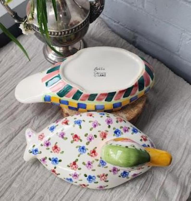 May include: Two decorative ceramic serving dishes. One dish is white with a floral pattern of pink, blue, and purple flowers. The other dish has a white base with colorful stripes and a checkered pattern. Both dishes have a unique shape.