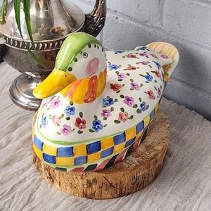 May include: A decorative ceramic duck-shaped container with a green head, yellow beak, and floral patterned body. The container is set on a wooden slice, with a checkered pattern around the base. The duck has pink cheeks and is painted with colorful flowers.
