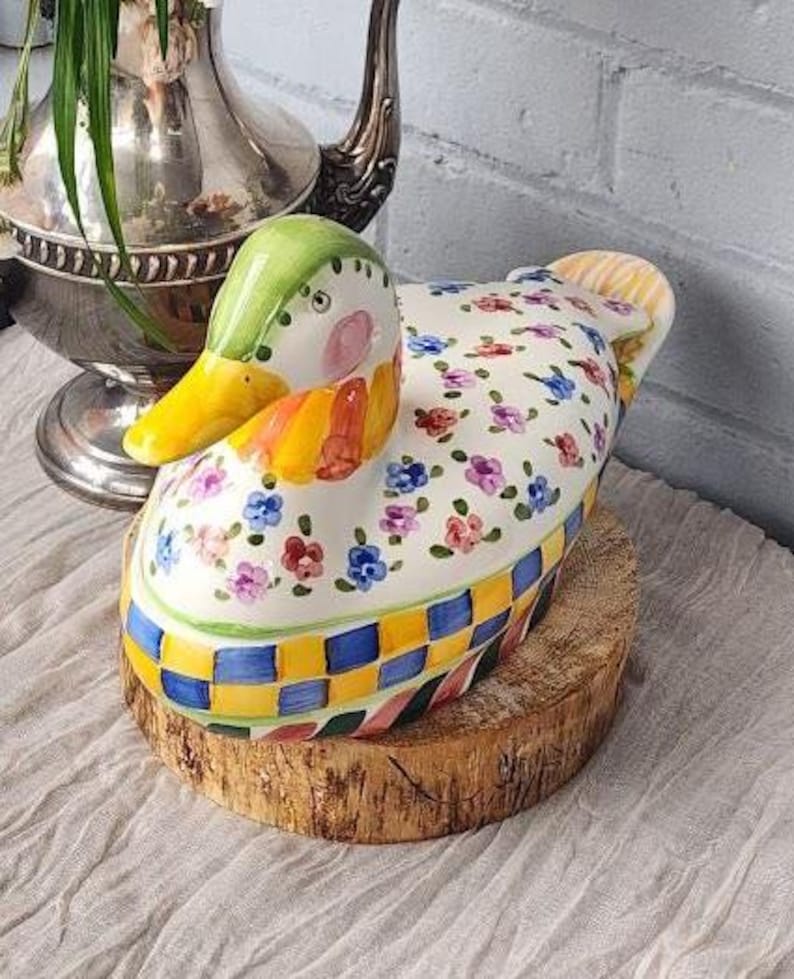 May include: A decorative ceramic duck-shaped container with a green head, yellow beak, and floral patterned body. The container is set on a wooden slice, with a checkered pattern around the base. The duck has pink cheeks and is painted with colorful flowers.