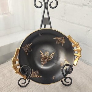 May include: A black bowl with gold-colored decorative handles and gold-colored line art patterns on the interior surface. The bowl is displayed on a black metal stand featuring scrollwork details at the base and a tall, curved support structure.