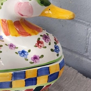 May include: A ceramic duck-shaped container with a green head, yellow beak, and pink cheeks. The body is white with floral and checkered patterns in various colors. The container is placed on a wooden base.