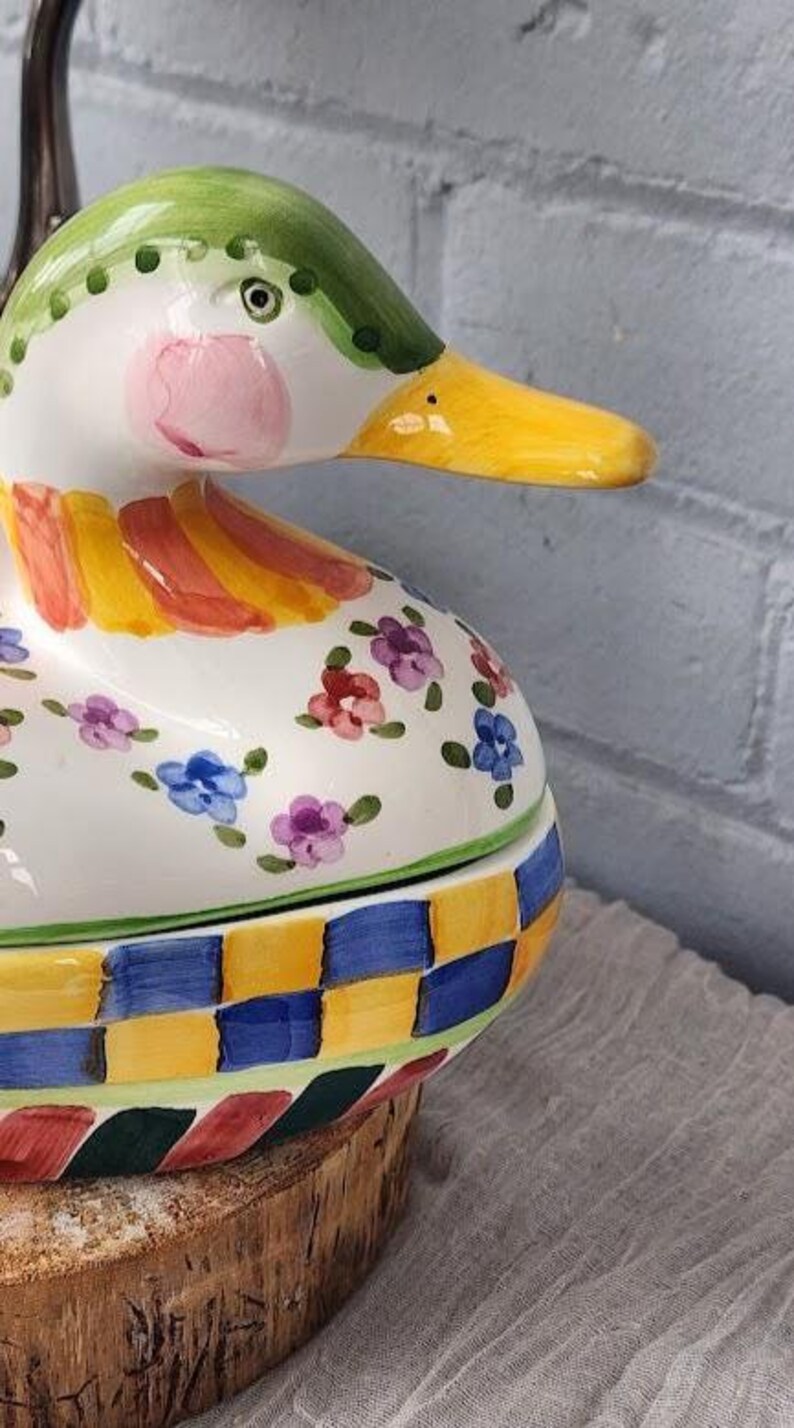 May include: A ceramic duck-shaped container with a green head, yellow beak, and pink cheeks. The body is white with floral and checkered patterns in various colors. The container is placed on a wooden base.
