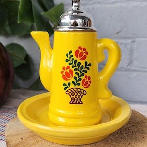 May include: A bright yellow ceramic teapot with a silver lid, set on a matching saucer. The teapot features a floral design in red, green, and black. The teapot is sitting on a wooden surface.
