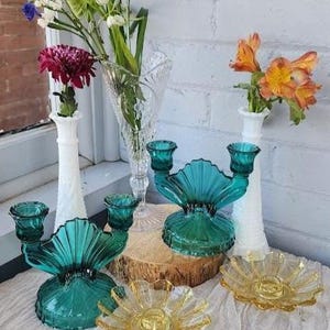 May include: Two teal glass double-candle holders with fan-shaped centers, two white milk glass bud vases with spiral textures, one clear pressed glass vase, and two yellow glass flower-shaped candle plates arranged on a white fabric surface.