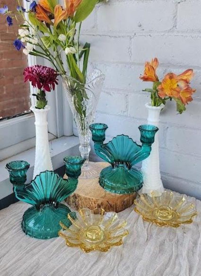 May include: Two teal glass double-candle holders with fan-shaped centers, two white milk glass bud vases with spiral textures, one clear pressed glass vase, and two yellow glass flower-shaped candle plates arranged on a white fabric surface.