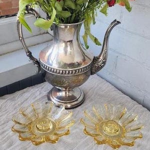 May include: A silver-plated teapot with an ornate handle and spout, filled with a bouquet of red, orange, and green flowers. Two clear, amber-tinted glass candle holders with a textured, petal-like design are positioned in front of the teapot.