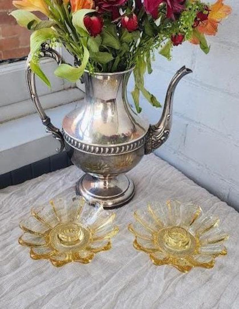 May include: A silver-plated teapot with an ornate handle and spout, filled with a bouquet of red, orange, and green flowers. Two clear, amber-tinted glass candle holders with a textured, petal-like design are positioned in front of the teapot.