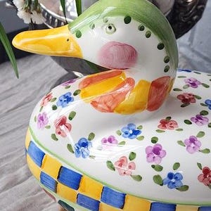 May include: A decorative ceramic duck-shaped container with a lid. The duck has a green head, yellow beak, and pink cheeks. The body is white with colorful floral and checkered patterns. The container sits on a wooden base.