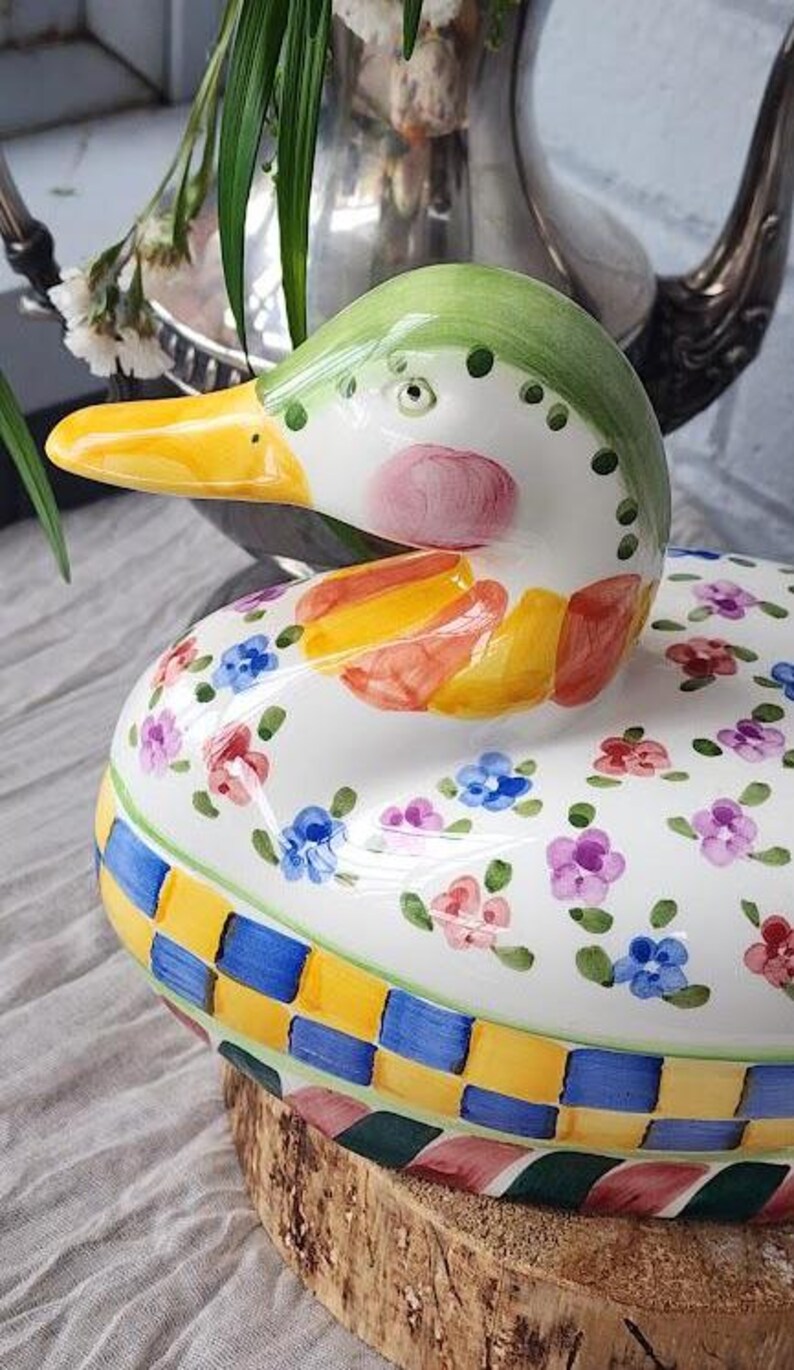 May include: A decorative ceramic duck-shaped container with a lid. The duck has a green head, yellow beak, and pink cheeks. The body is white with colorful floral and checkered patterns. The container sits on a wooden base.