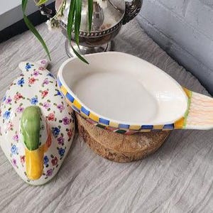 May include: A decorative ceramic duck-shaped dish and a matching oval serving dish. The duck dish is white with floral patterns and a green and yellow beak. The serving dish has a checkered rim and rests on a wooden base.