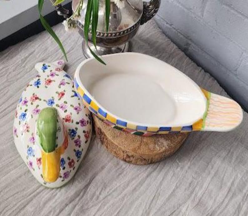 May include: A decorative ceramic duck-shaped dish and a matching oval serving dish. The duck dish is white with floral patterns and a green and yellow beak. The serving dish has a checkered rim and rests on a wooden base.