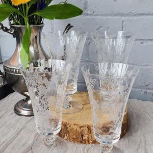 May include: A collection of clear, etched glass goblets with floral designs. The goblets have a flared shape and sit on a round base. A silver vase with flowers is in the background. The goblets are on a wooden surface.
