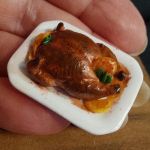 Peut inclure: Miniature poulet rôti sur une assiette blanche. Le poulet est de couleur brun foncé, garni de tranches d'orange et d'herbes vertes. L'assiette est carrée avec des coins arrondis.